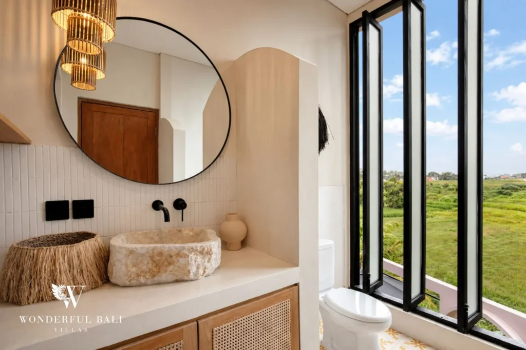 An en-suite bathroom with a stone basin, round mirror and window over the rice fields at a 3-bedroom villa in Pererenan, Bali