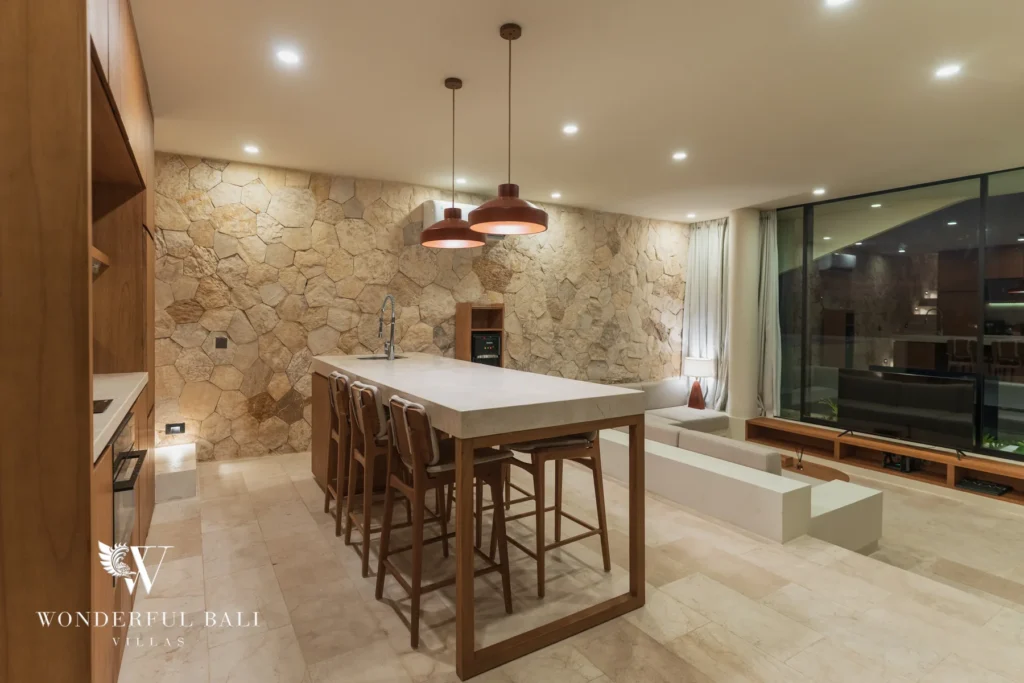 A kitchen and dining area with a stone island, bar stools, copper pendant lights and a natural stone wall in the evening at a 3-bedroom villa in Bingin, Uluwatu, Bali