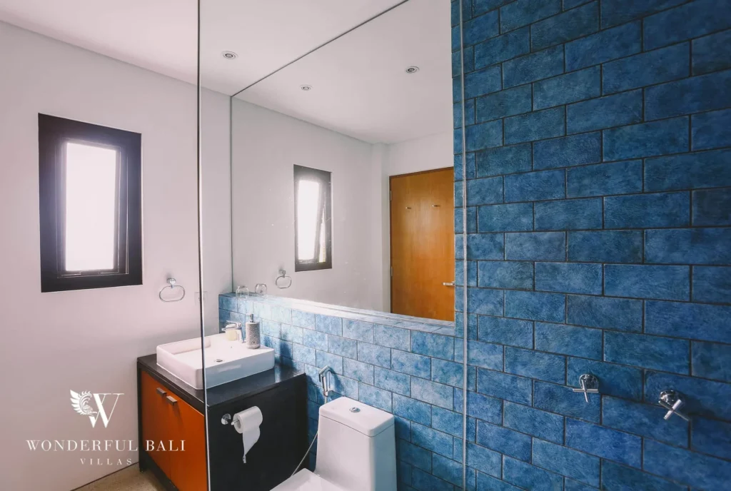 A bathroom with blue-tiled walls, a large mirror, a wall-mounted basin and a toilet beside a slim window at a 4-bedroom villa in Batu Bolong, Canggu, Bali