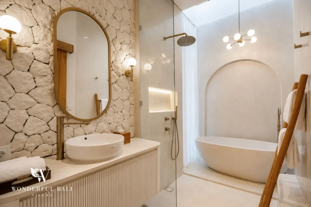 A stone-walled bathroom with a fluted vanity, oval mirror, glass shower and a freestanding tub under a globe chandelier, at a 2-bedroom villa in Bingin, Uluwatu, Bali
