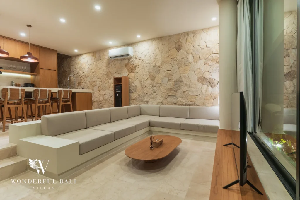 An evening living room with an L-shaped sofa, low timber coffee table and a stone feature wall beside the open kitchen at a 3-bedroom villa in Bingin, Uluwatu, Bali
