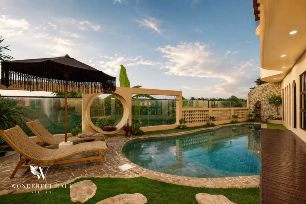 The pool, loungers and fringed umbrella beside the stone-clad villa wall at twilight at a 3-bedroom villa in Pererenan, Bali