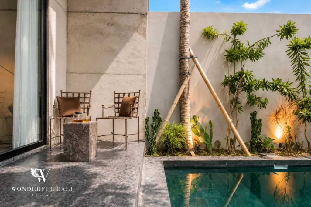 Sun-lit terrace corner with two metal chairs, a stone side table and a palm beside the pool against a concrete wall at a 2-bedroom villa in Seseh, Bali