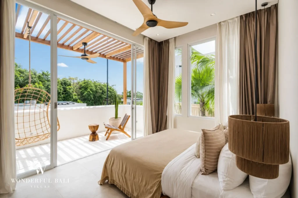 A bedroom with a beige bed opening onto a balcony with a hanging rattan chair and treetop views, at a 2-bedroom villa in Bingin, Uluwatu, Bali