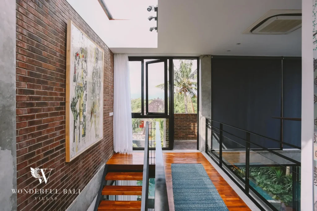 An upstairs landing with a red-brick wall, framed map artwork and a glass door opening to a balcony with treetop views at a 4-bedroom villa in Batu Bolong, Canggu, Bali