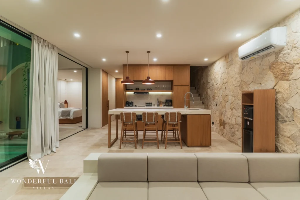 An open-plan living and kitchen space with a stone wall, island bar and a bedroom visible through glass doors at a 3-bedroom villa in Bingin, Uluwatu, Bali
