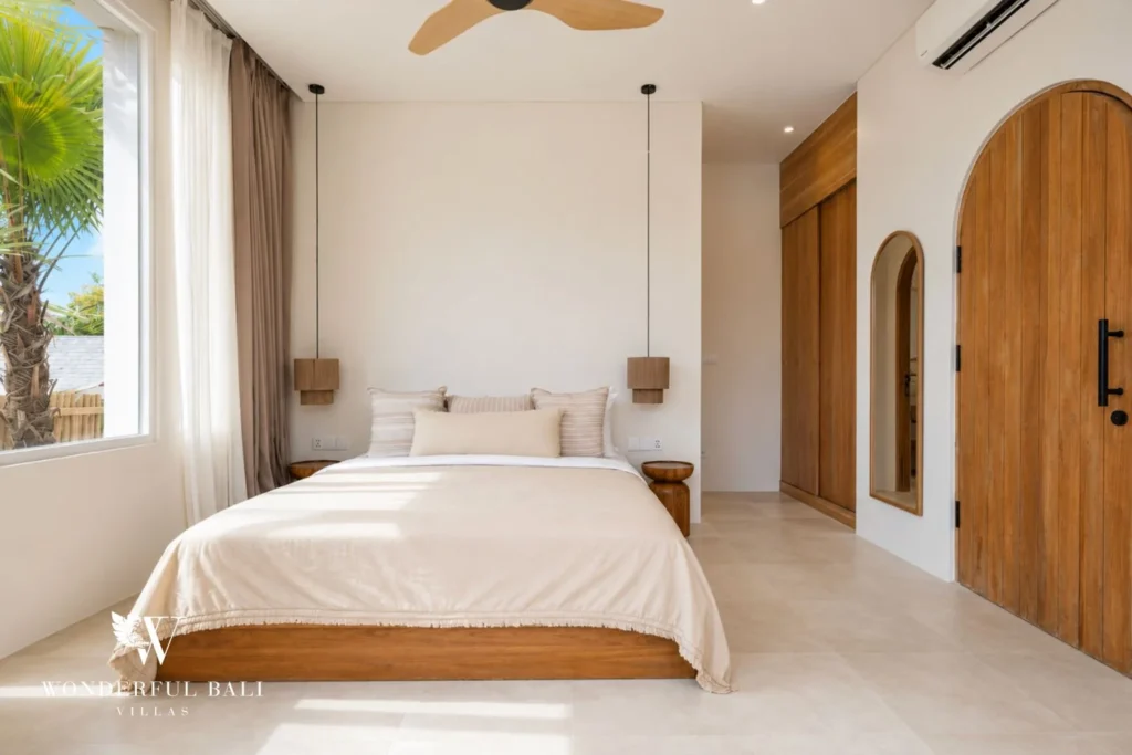 A bedroom with a wooden platform bed, pendant nightstands and an arched timber door beside a bright window, at a 2-bedroom villa in Bingin, Uluwatu, Bali