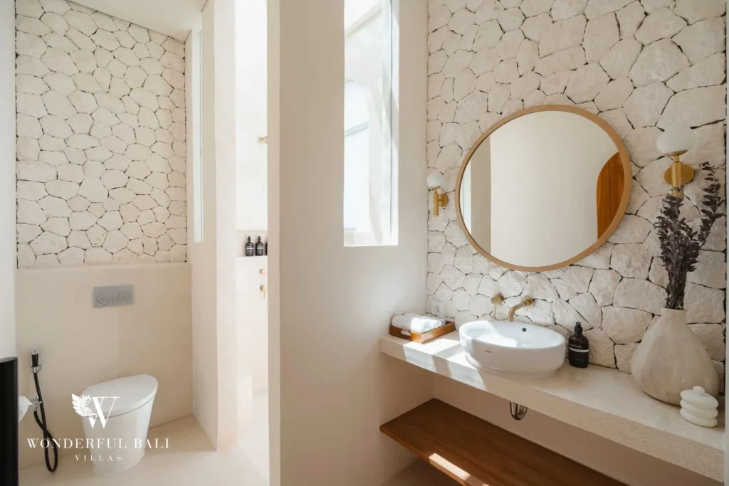 A powder room with a round mirror, stone-clad wall, wall-mounted basin and a separate toilet nook, at a 2-bedroom villa in Bingin, Uluwatu, Bali