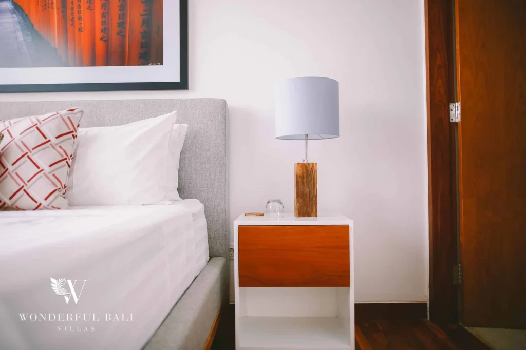 A bedside detail with a grey headboard, white bedding, a timber-fronted nightstand and a wood-based lamp at a 4-bedroom villa in Batu Bolong, Canggu, Bali