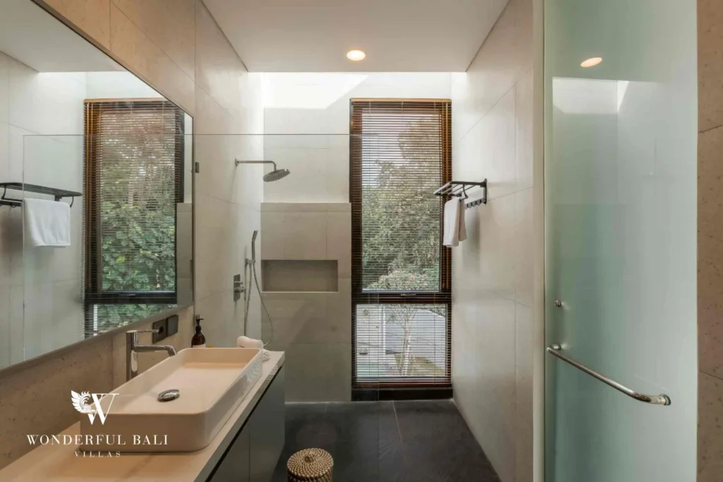 A stone-tiled ensuite with a walk-in rain shower and vessel sink framed by garden-view windows at a 4-bedroom villa in Pererenan, Bali
