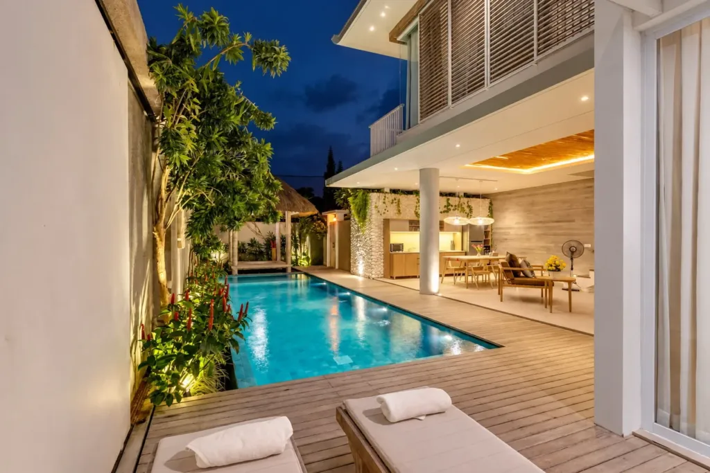 Villa Swan 02 — pool, sun loungers on the timber deck beside the lit water with the living room glowing at dusk