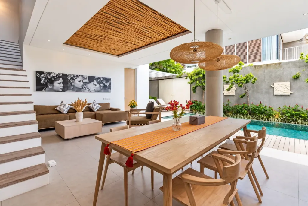Villa Swan 02 — dining, long timber table for eight under woven rattan pendants, living room beyond