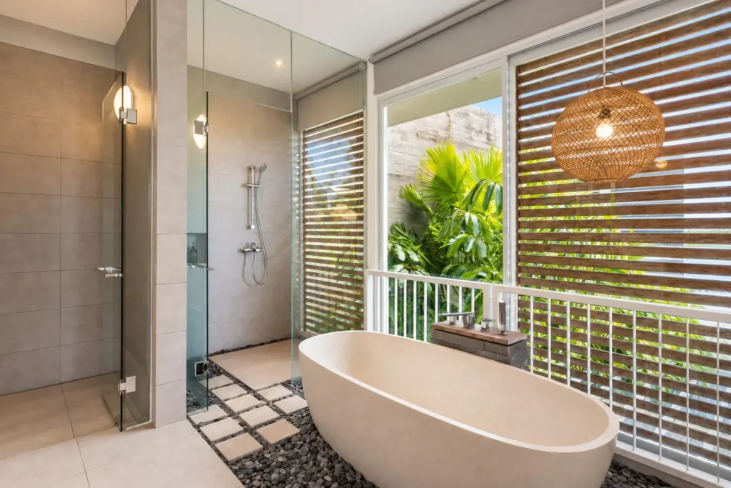 Villa Swan 02 — bathroom 1, freestanding bathtub and glass rain shower beside a louvred window onto tropical planting