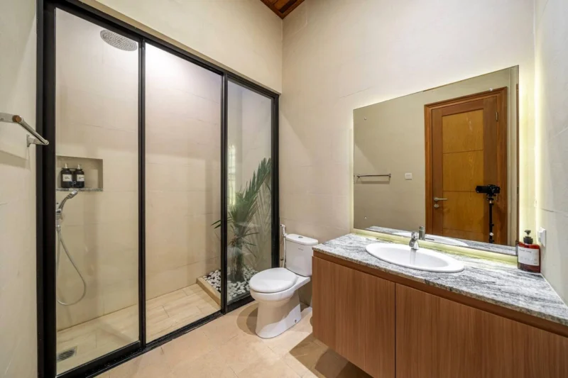 Villa LaBumbak, Umalas — bathroom 1, walk-in rain shower behind black-framed glass, marble-topped vanity with a backlit mirror, planting and pebbles behind the screen