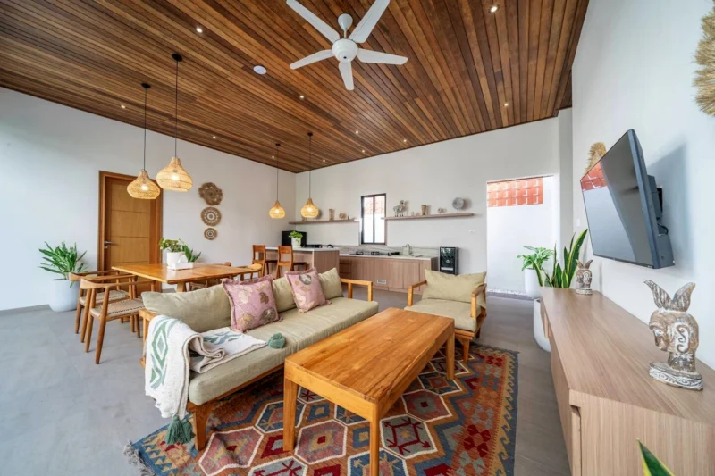 Villa LaBumbak, Umalas — living, sofa and coffee table under a dark timber ceiling, wall TV, dining table and kitchen beyond