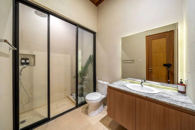 Villa LaBumbak, Umalas — bathroom 1, walk-in rain shower behind black-framed glass, marble-topped vanity with a backlit mirror, planting and pebbles behind the screen