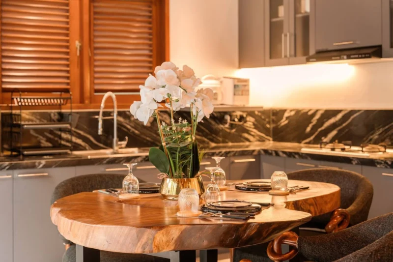 Dining table close-up in Seminyak, Bali — white orchid in gold vase, wine glasses, black ceramic plates on live-edge suar wood grain, dark marble splashback kitchen behind.
