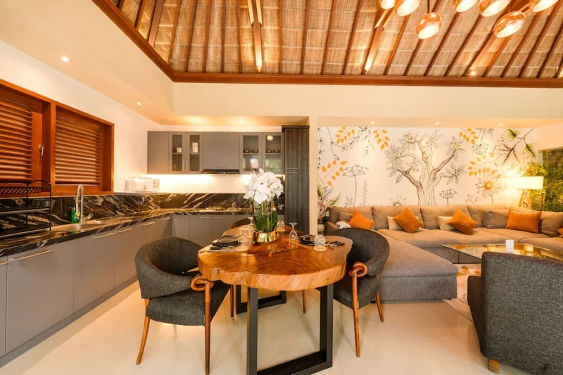 Live-edge suar wood dining table detail in Seminyak, Bali — set for two with wine glasses, black ceramic plates, white orchid in gold vase, dark marble splashback kitchen behind.