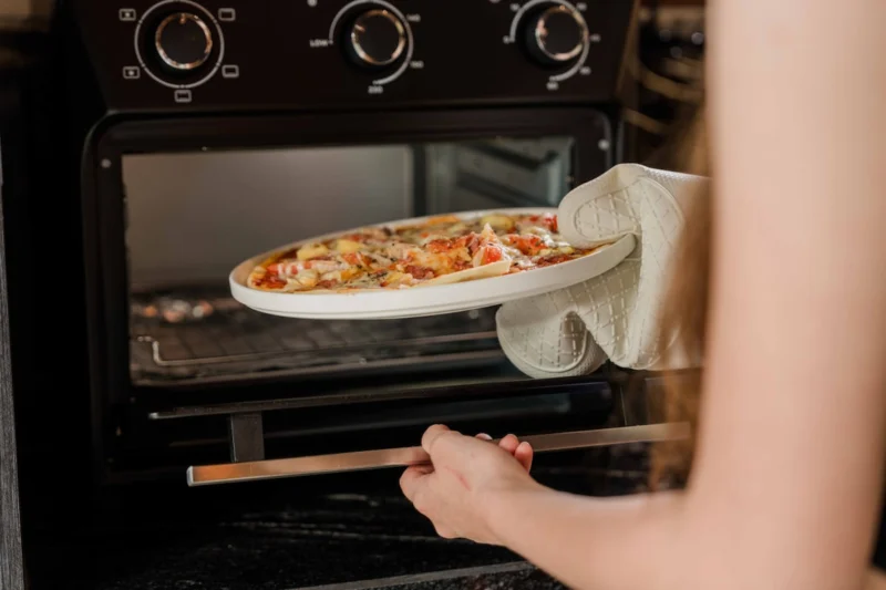 Homemade pizza being removed from kitchen oven in Seminyak, Bali — freshly baked pizza on white ceramic plate, oven gloves, dark marble splashback visible behind.