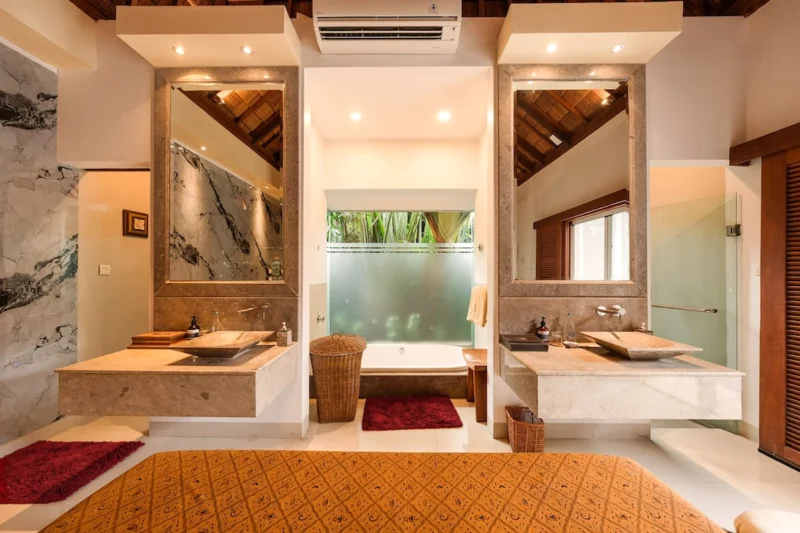 Symmetrical master bathroom in Seminyak, Bali — dual travertine vanities with vessel sinks, central soaking tub behind frosted glass, marble wall, thatched ceiling in both mirrors.
