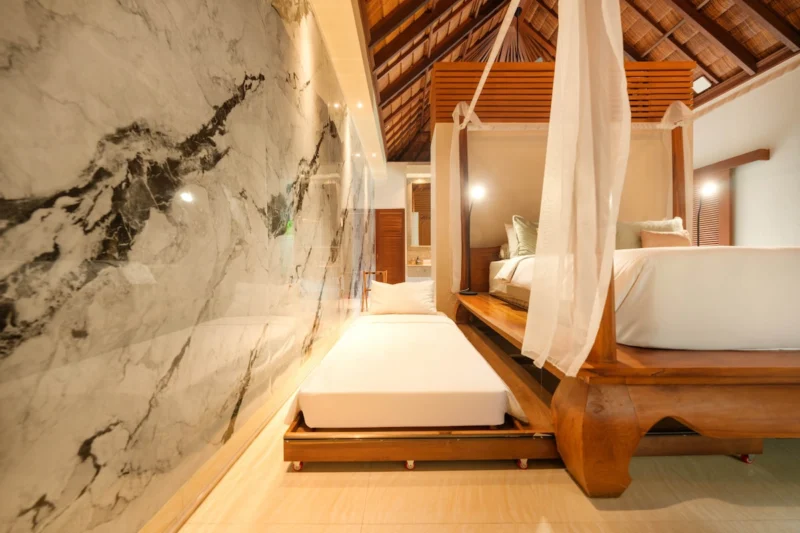 Master bedroom in Seminyak, Bali — four-poster canopy bed with trundle extended below, floor-to-ceiling marble feature wall with uplighting, ensuite spa bathroom visible through right opening.