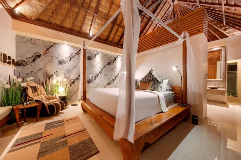 Bedroom bed detail in Seminyak, Bali — king bed with batik print and sage cushions, white linen, marble feature wall behind headboard, garden window with tropical greenery left.