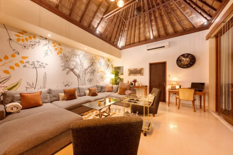 Living room in Seminyak, Bali — large sectional sofa with terracotta cushions, botanical tree mural, gold bar trolley, dedicated desk and chair workspace corner right, alang-alang ceiling.