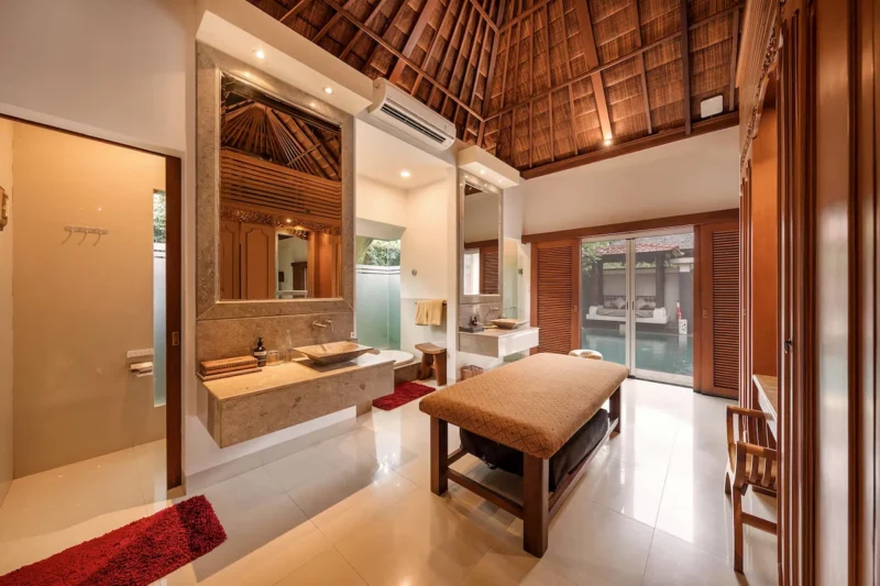 Full spa bathroom layout in Seminyak, Bali — double floating travertine vanity with vessel sinks, massage table, frosted glass tub enclosure, thatched ceiling, glass doors to pool beyond.