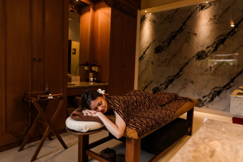Guest resting on massage table in Seminyak, Bali — traditional batik fabric draped, frangipani in hair, marble feature wall behind, carved Balinese wardrobe lit warmly in background.