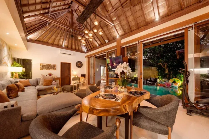 Open-plan living and dining room in Seminyak, Bali — live-edge suar wood dining table, sectional sofa, copper pendant cluster, full-width glass doors framing private pool and tropical garden.