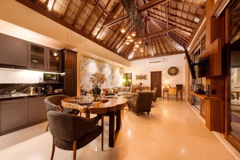Open-plan kitchen through dining to living room in Seminyak, Bali — dark marble kitchen, live-edge dining table, sectional sofa, botanical mural, copper pendants, Nespresso on teak credenza right.