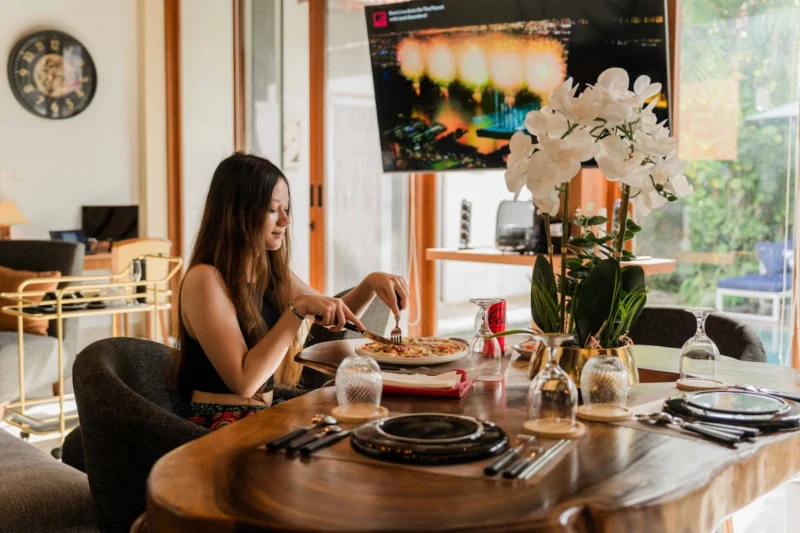 Dining lifestyle in Seminyak, Bali — woman eating at live-edge suar wood dining table, pool and tropical garden visible through glass sliding doors behind. Natural daytime light.