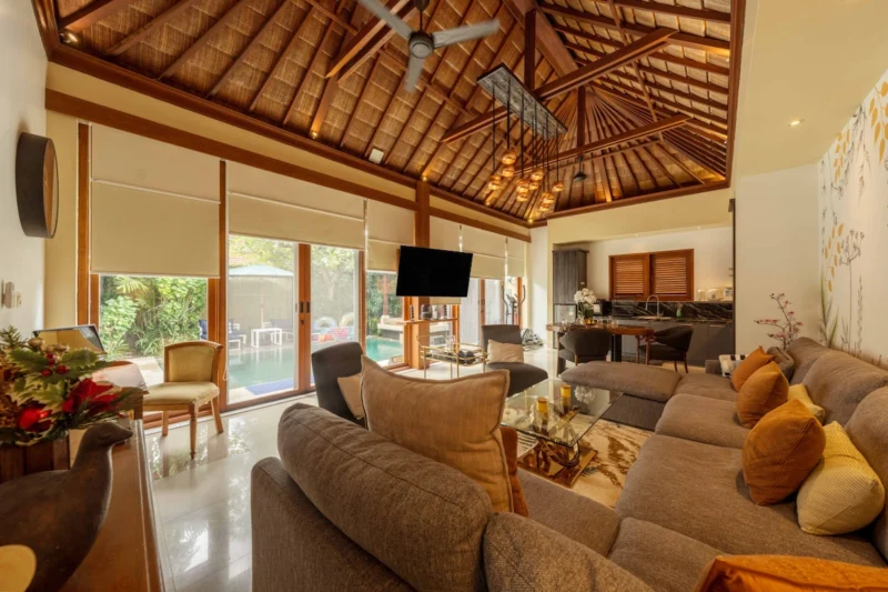 Daytime living room in Seminyak, Bali — sectional sofa with amber cushions, copper pendant cluster, alang-alang thatched ceiling, glass doors partially open to pool and tropical garden.