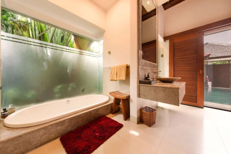 Deep soaking bathtub in Seminyak, Bali — travertine stone surround, frosted glass panel with tropical garden behind, stone vessel sink on floating vanity, louvred door to private pool right.