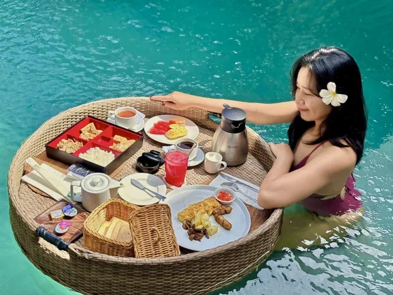 Floating breakfast service in Seminyak, Bali — woman with frangipani in private pool with large round rattan floating tray loaded with nasi goreng, eggs, toast, fruit, juice and coffee.