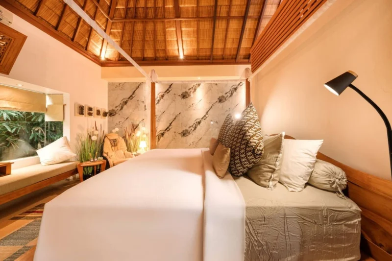 Master bedroom in Seminyak, Bali — king four-poster teak canopy bed, marble feature wall with spotlights, full-body massage chair with pampas grass, ensuite spa bathroom visible right.