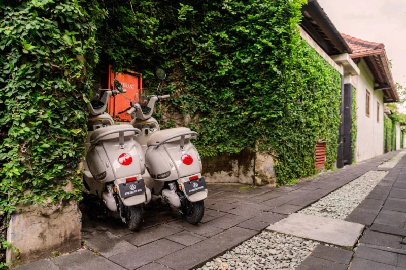 Two complimentary cream Pacific scooters in the villa laneway, Seminyak, Bali — parked against ivy-covered wall, stone paving, terracotta roof and tropical greenery beyond.