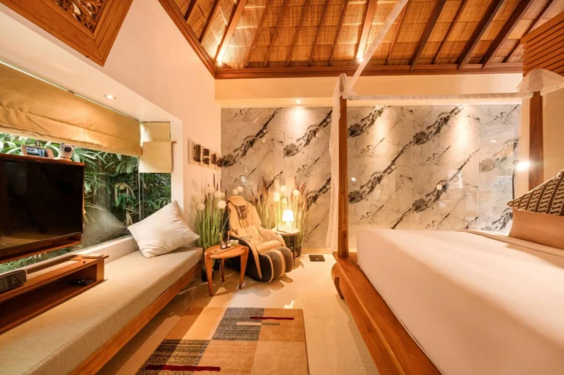 Full master bedroom overview in Seminyak, Bali — teak TV unit with garden window, built-in day bench, full-body massage chair against marble feature wall, king four-poster canopy bed right.