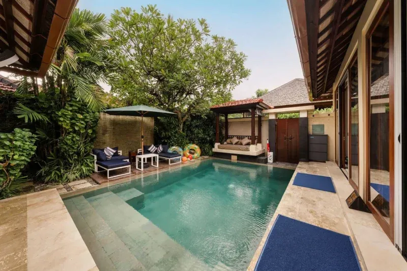 Private pool in Seminyak, Bali — navy sunloungers, Balinese bale daybed pavilion, tropical garden with frangipani tree, bamboo privacy screening, terracotta roof tiles, glass sliding doors.