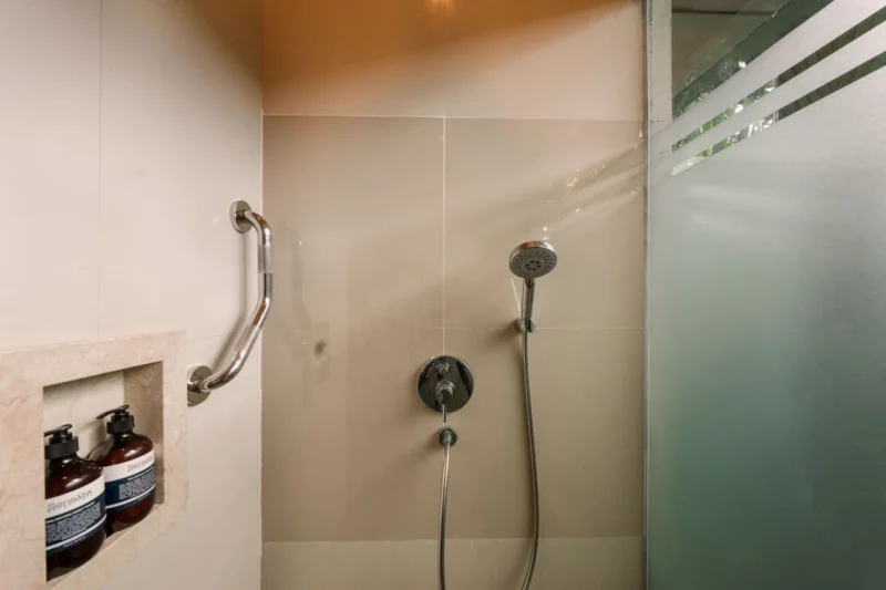 Walk-in shower in Seminyak, Bali — chrome handset shower, travertine stone niche with Sensatia Botanicals amenities, frosted glass partition, stone tile surround.