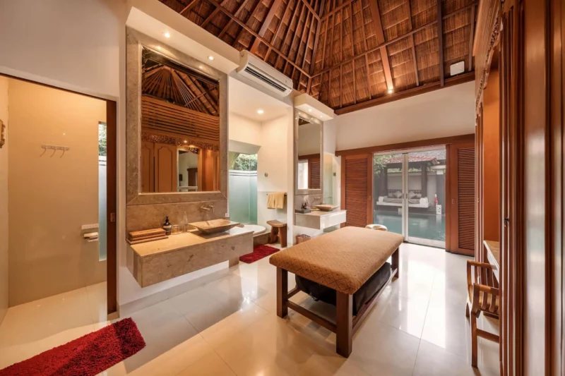 Five-star layout, entirely yours. The master bathroom at Villa 23 gives you a double stone vessel vanity, deep soaking tub behind frosted garden glass, dedicated massage table, and a direct view through to the private pool — a complete wellness suite.