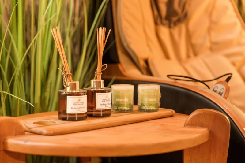 Wellness detail in Seminyak, Bali — two Bedugul Wood luxury reed diffusers and citronella candles on teak armrest tray of full-body massage chair, pampas grass softly behind.