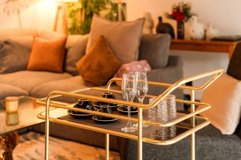 Gold bar trolley detail in Seminyak, Bali — two champagne flutes and glassware on gold and black two-tier bar cart, sectional sofa softly focused behind, warm ambient lighting.