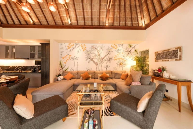 Living room in Seminyak, Bali — large sectional sofa facing full-wall botanical tree mural, gold bar trolley with champagne flutes, glass coffee table on botanical rug, kitchen visible left.