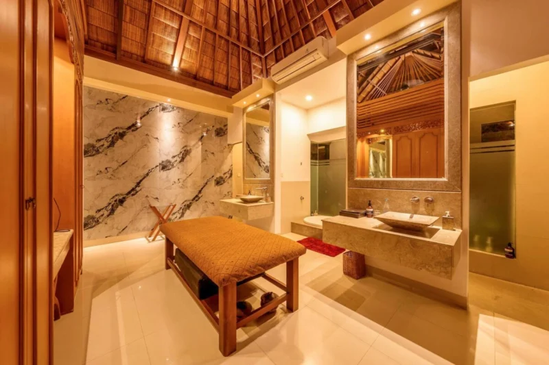 Spa bathroom in Seminyak, Bali — floor-to-ceiling marble feature wall with bold dark veining, massage table foreground, double stone vessel sinks on travertine vanity, thatched ceiling in ornate stone mirror.