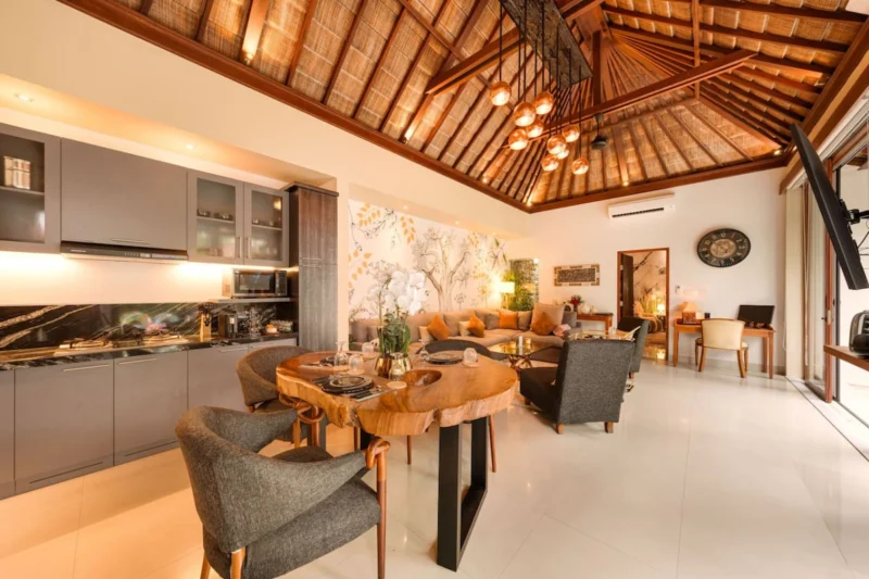Open-plan kitchen and dining in Seminyak, Bali — live-edge suar wood dining table, dark marble splashback kitchen, copper pendant cluster, botanical mural, alang-alang thatched ceiling.