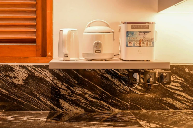 Kitchen appliance shelf in Seminyak, Bali — electric kettle, rice cooker, and fast ice maker on white shelf above dark marble splashback, teak louvred shutters behind.