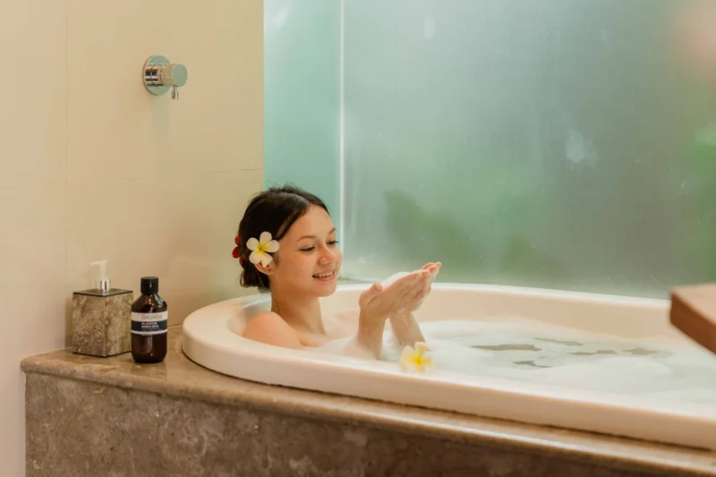 Bathtub lifestyle in Seminyak, Bali — smiling woman with frangipani in hair in deep soaking tub, Sensatia Botanicals Relaxation Bubble Bath on travertine surround, frosted glass behind.