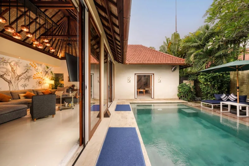 Indoor-outdoor connection in Seminyak, Bali — view from living room threshold across travertine pool terrace to private pool, navy sunloungers, copper pendants and botanical mural visible inside.