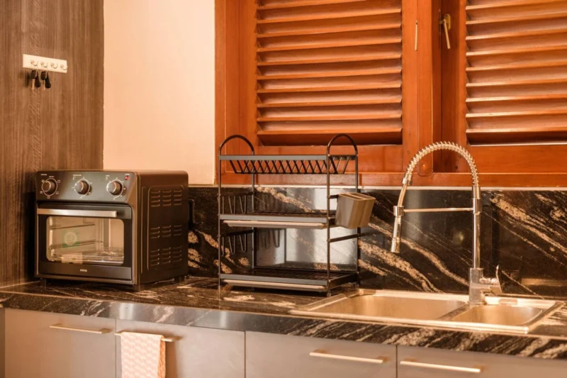 Kitchen sink end in Seminyak, Bali — countertop oven, double stainless sink with chrome mixer tap, dark marble splashback with gold veining, grey cabinetry, teak louvred shutters.